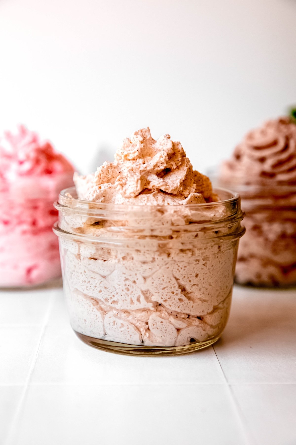 small glass jar of cinnamon whipped cream from a dispenser on a white table with a small jar of strawberry whipped cream and chocolate whipped cream blurred out on either side in the background.