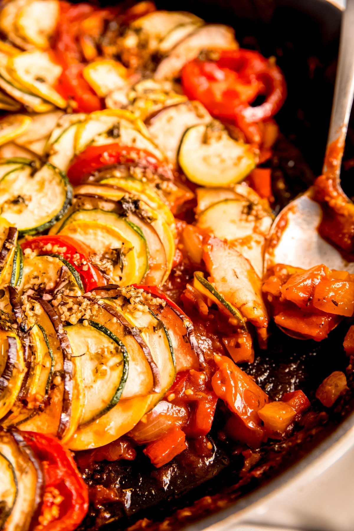closeup 45 degree angle shot of a disney's ratatouille (confit biyaldi) with a silver spoon taking a serving out showing the thinly sliced veggies atop the chunky tomato pepper and onion sauce.