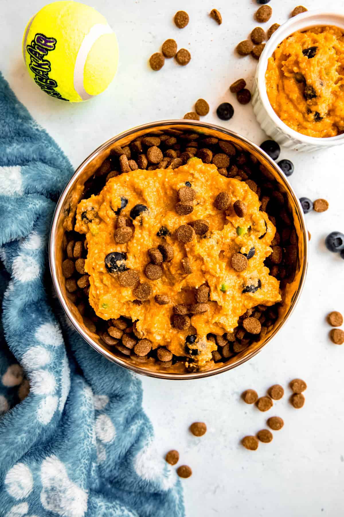 flat lay shot of a dog's bowl filled with dry kibble topped with homemade pumpkin, chicken, and rice dog food topper on a white table with a blue dog blanket, a dog's tennis ball, and blueberries and kibble scattered around.