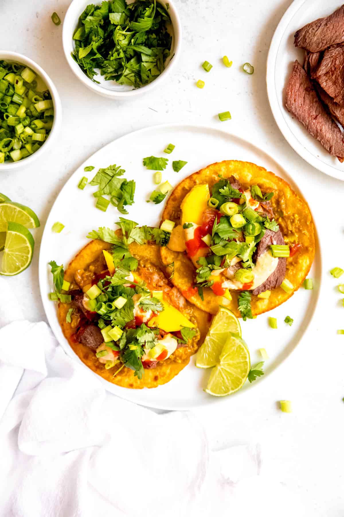 overhead shot of a white dinner plate with two grilled skirt steak tacos with mango-gochujang guacamole, scallions, cilantro, and yumyum sauce on a white table with bowls of extra lime wedges, green onions, chopped cilantro, and a plate of extra Korean BBQ beef.