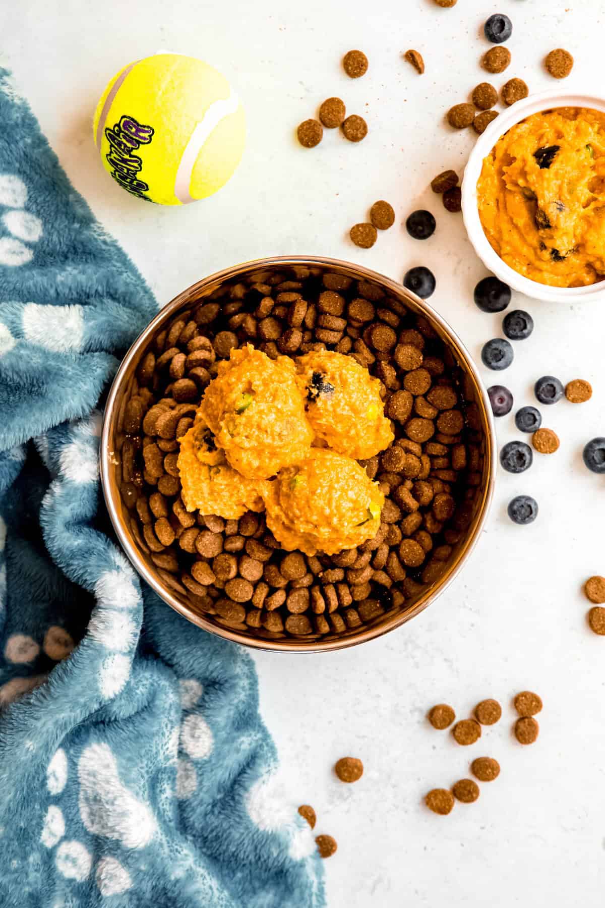 several scoops of DIY dog food topper added to a bowl of dry kibble on a white table with a dog's ball and snuggle blanket.