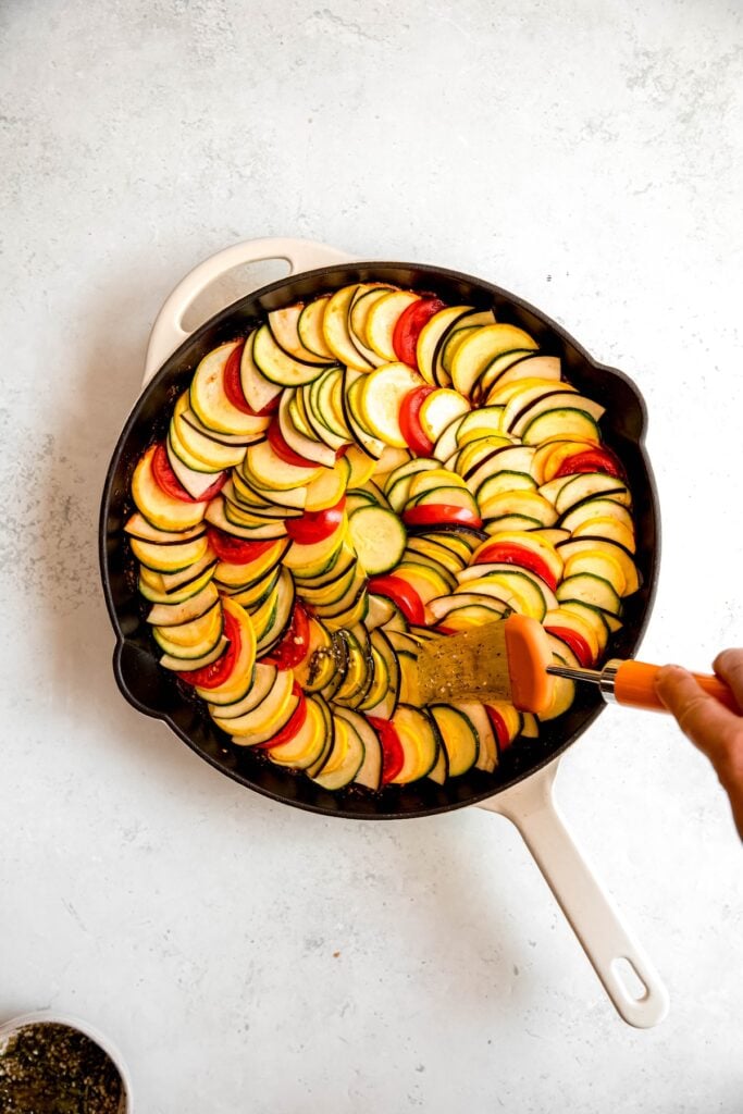 action shot of a hand brushing the tops of the ratatouille vegetables with garlic herb olive oil before baking.