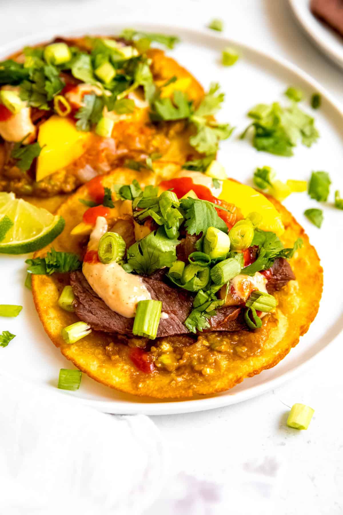 45 degree angle shot of a white plate with two crispy skirt steak asian tacos with Korean BBQ beef, spicy mango guacamole, yum yum sauce, gochujang, and yum yum sauce.