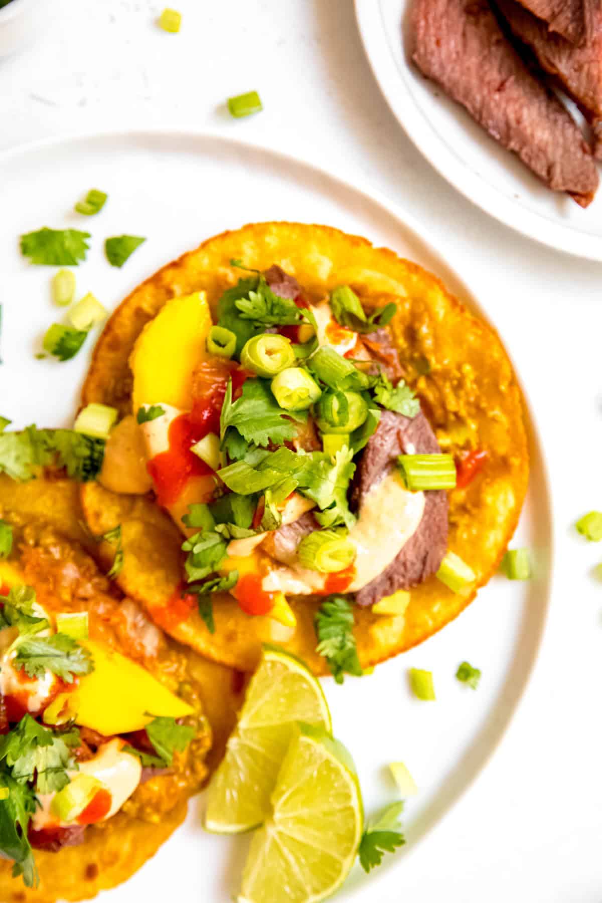 skirt steak korean tacos on a white plate with two lime wedges and chopped scallions and cilantro scattered around.