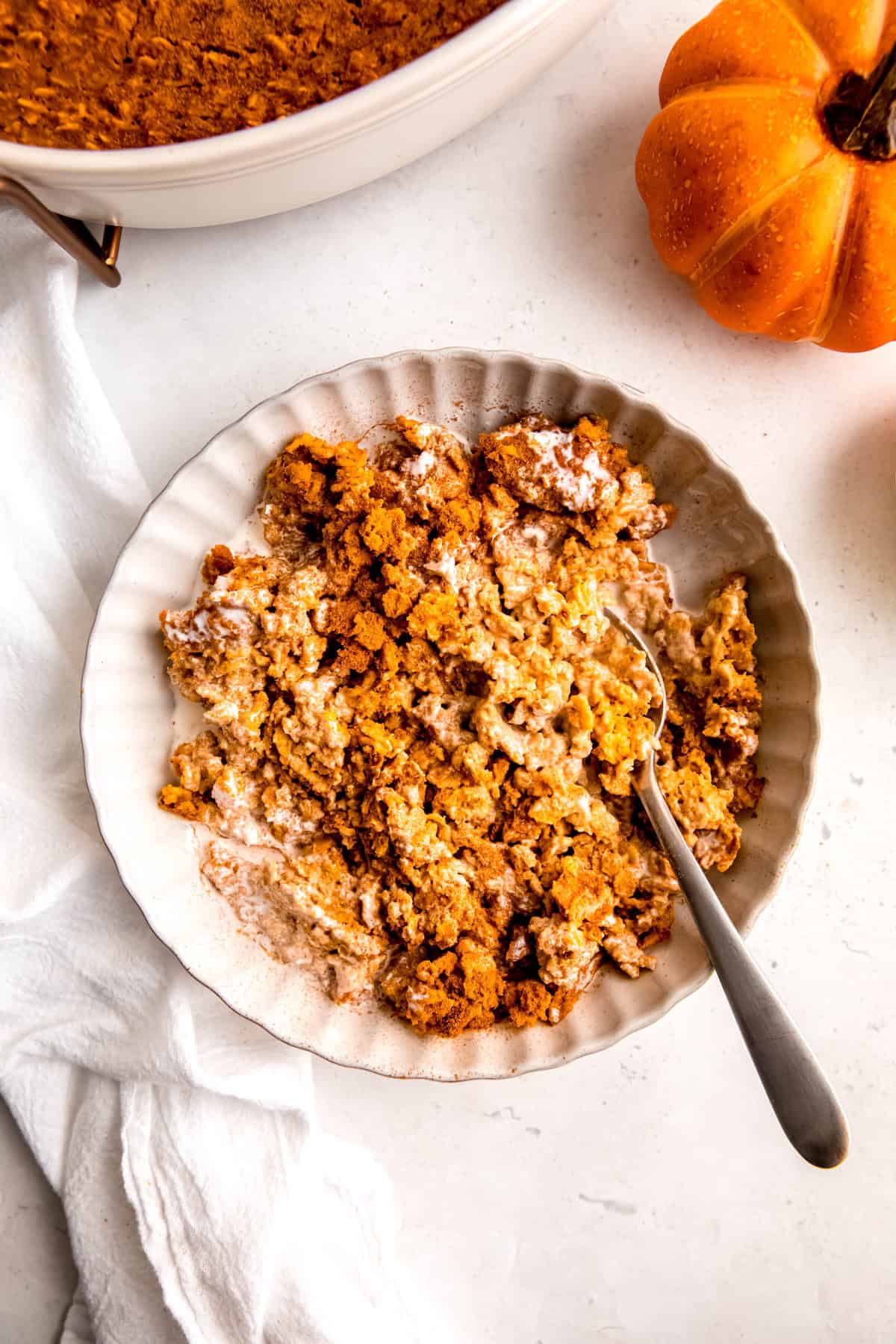 flat lay shot of a helping of baked pumpkin pie oatmeal in a white bowl on a white table with a mini pumpkin and the edge of the casserole dish with the remaining oat bake framing the top half of the shot.