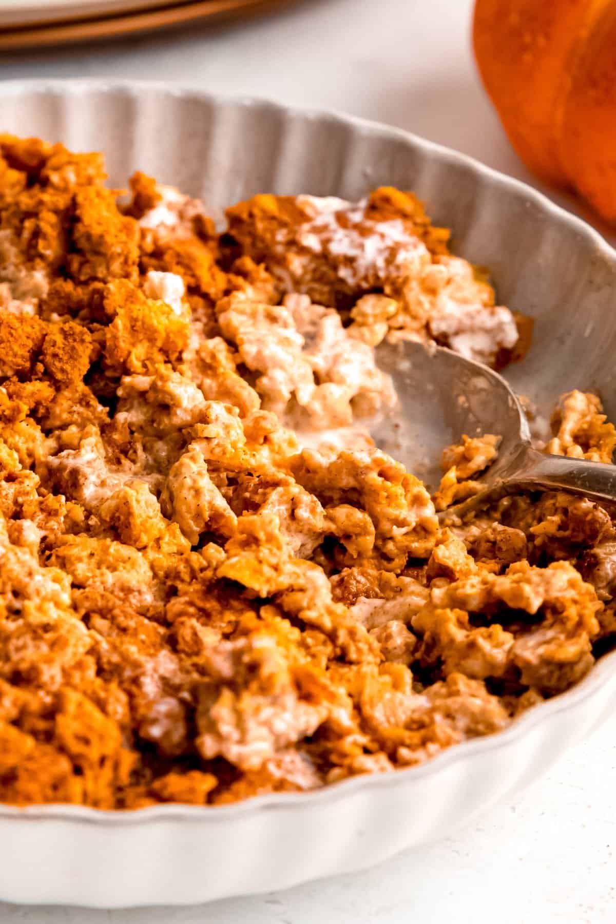 closeup shot of a serving of baked pumpkin pie oats in a scalloped white bowl topped with heavy cream and a light dusting of cinnamon with a silver spoon digging in for the next bite.