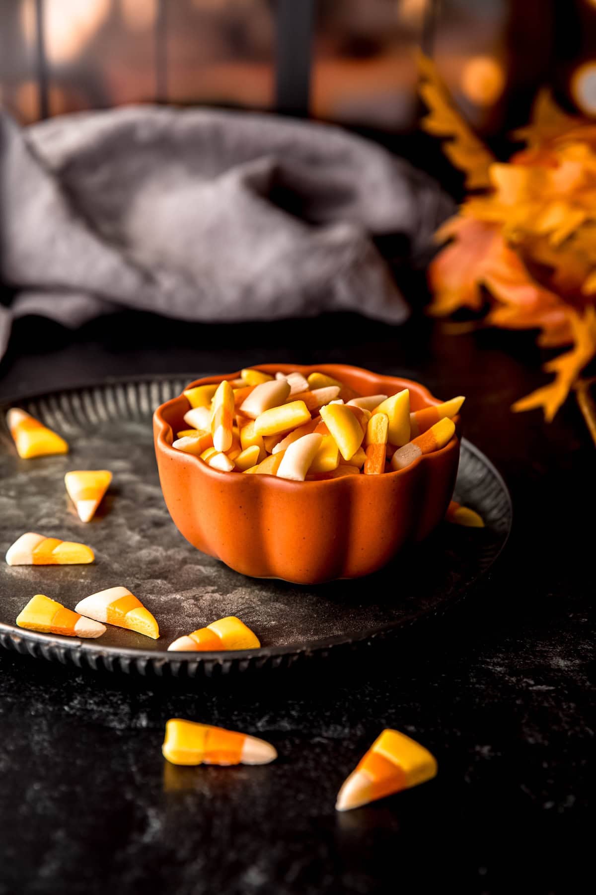 Halloween candy corn in a pumpkin shaped bowl on a black metal tray on a black table in front of a row of windows with black dividers and light coming through.