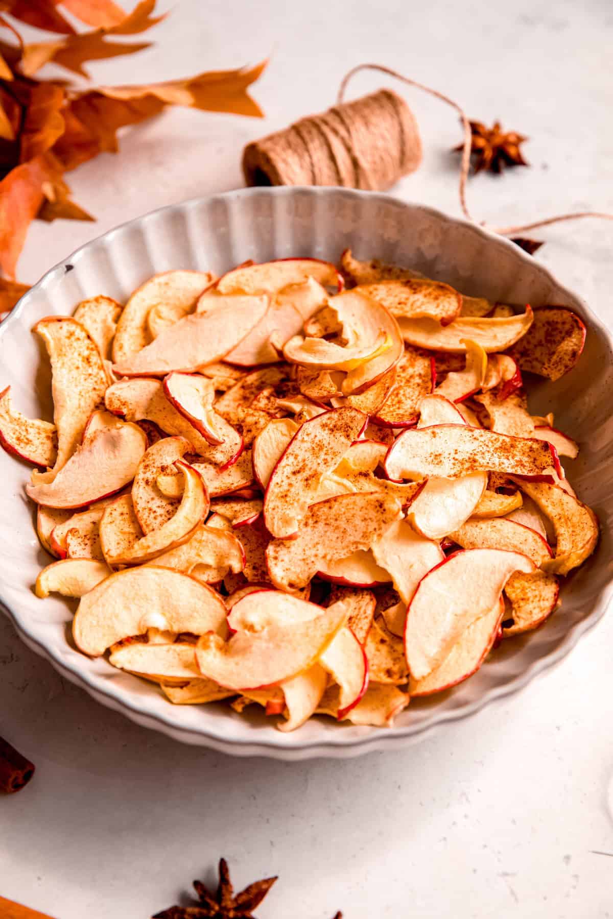 white scalloped serving bowl filled with crispy apple cinnamon chips on a white table with fall leaves, a few whole star anise, and a roll of natural twine.