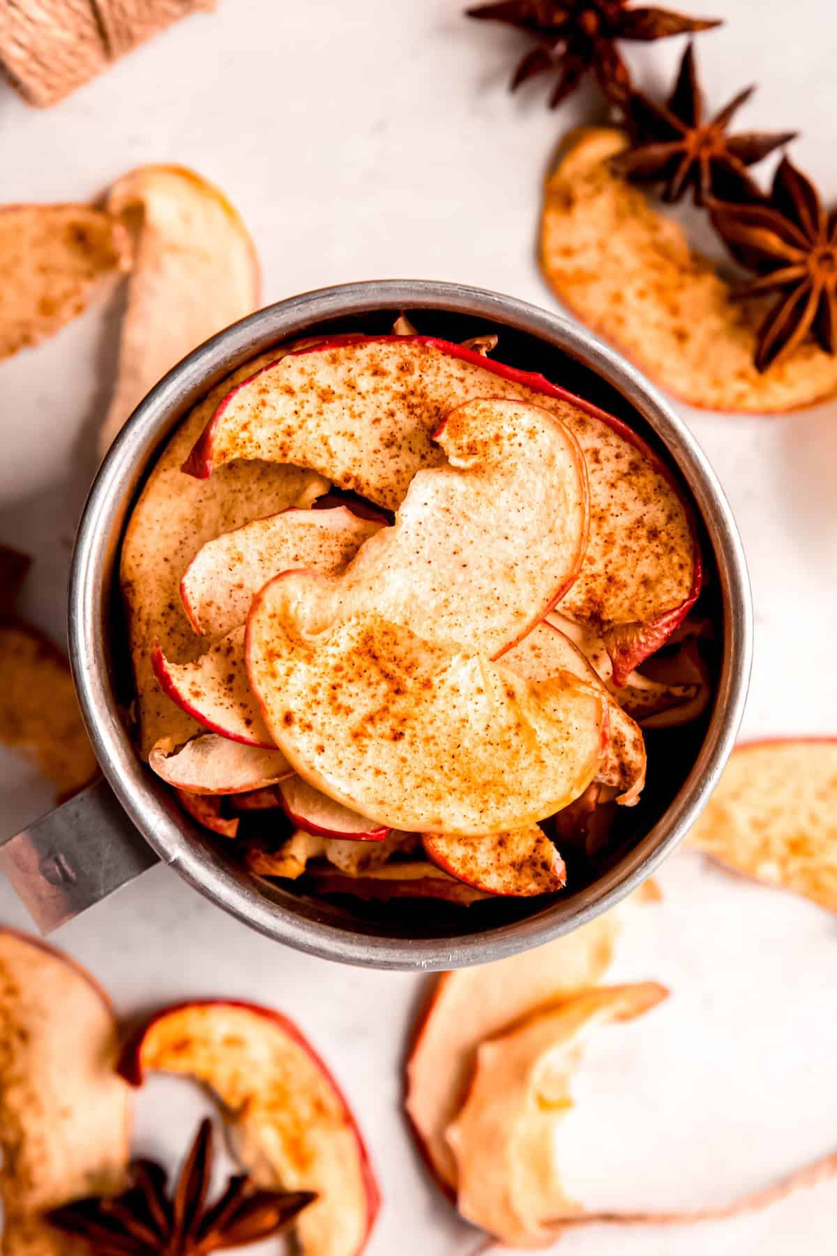 overhead shot of a silver tiffin of dehydrated apple chips on a white table with mrore apple chips and whole star anise blurred out on the table below.