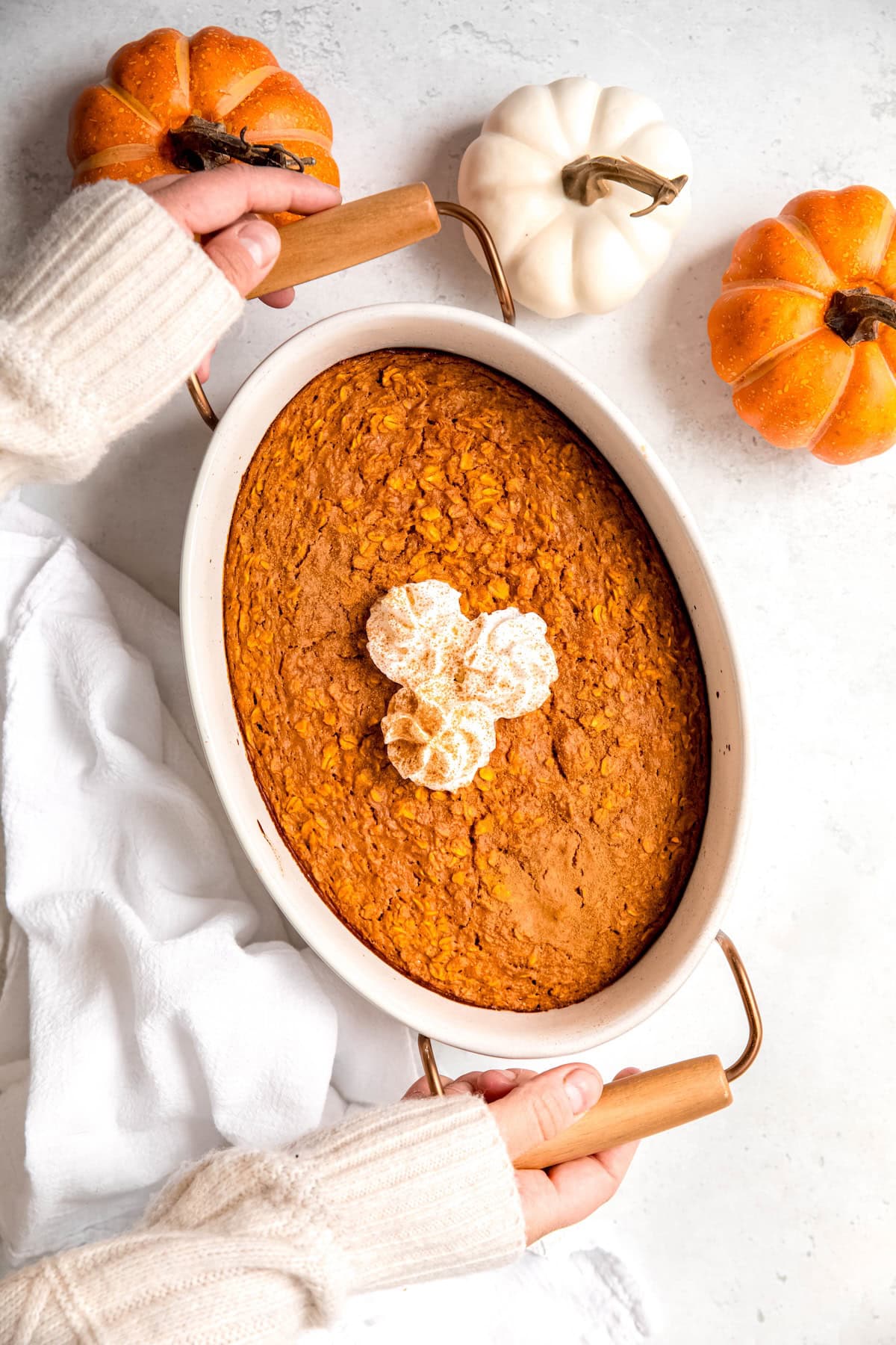 hands setting down a round white casserole dish filled with easy pumpkin baked oatmeal topped with a few squirts of whipped cream and a sprinkle of pumpkin spice down on a white table with 3 mini pumpkins.