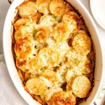 closeup shot of gratin potatoes with fennel in a white oval serving dish with rose gold and wood handles.