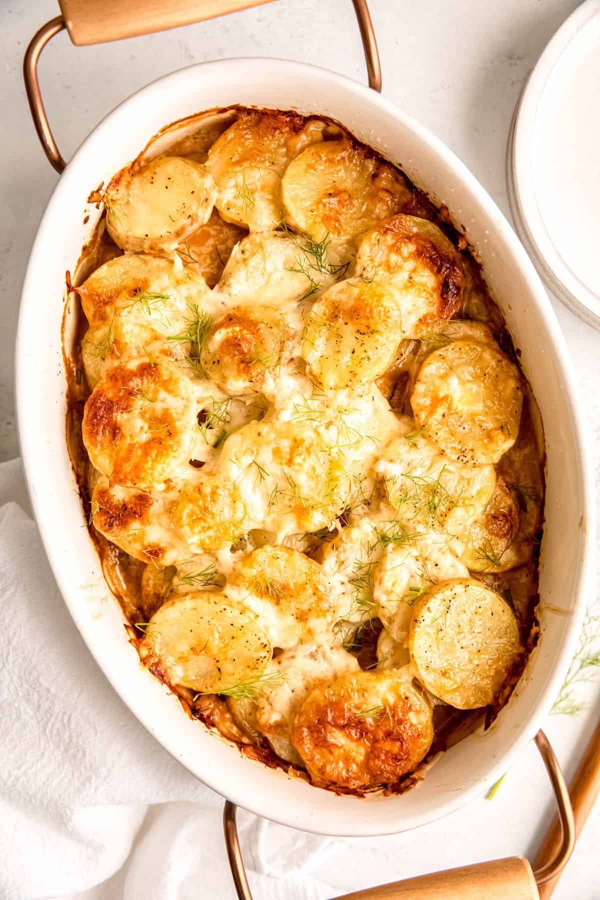 closeup shot of gratin potatoes with fennel in a white oval serving dish with rose gold and wood handles.