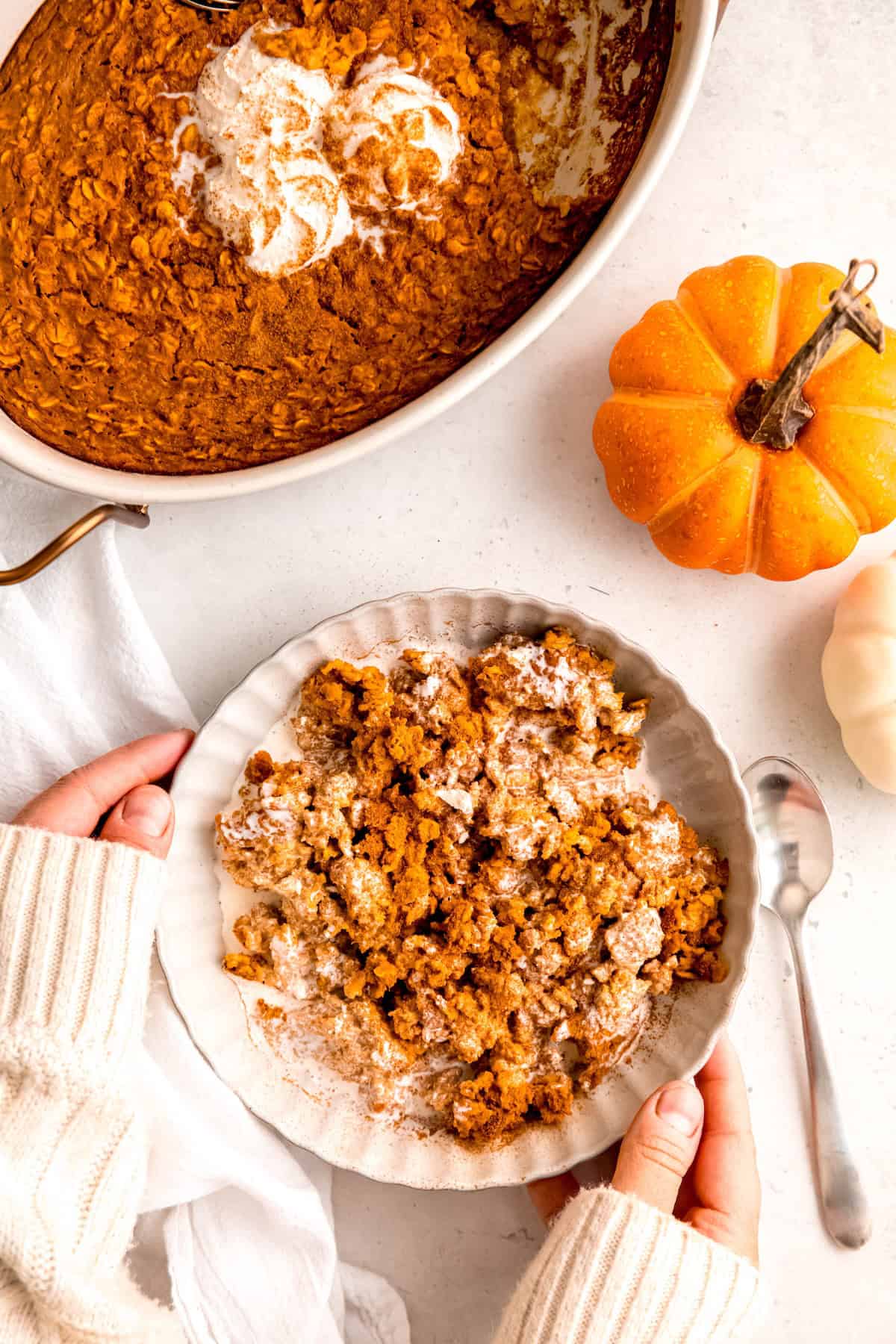 two hands in a white sweater serving a bowl of healthy pumpkin baked oatmeal on a white table next to a silver spoon with a few mini pumpkins and the remaining pumpkin spice baked oatmeal in the top half of the frame.