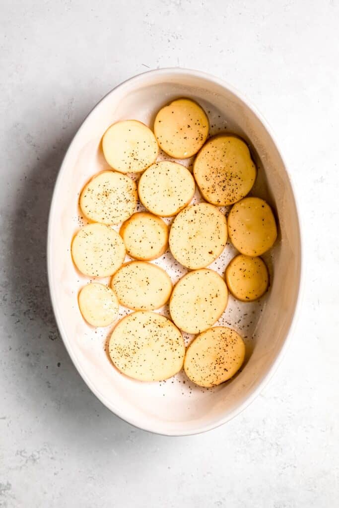 thinly sliced potatoes layered in the bottom of an oval white casserole dish and sprinkled with salt and black pepper.