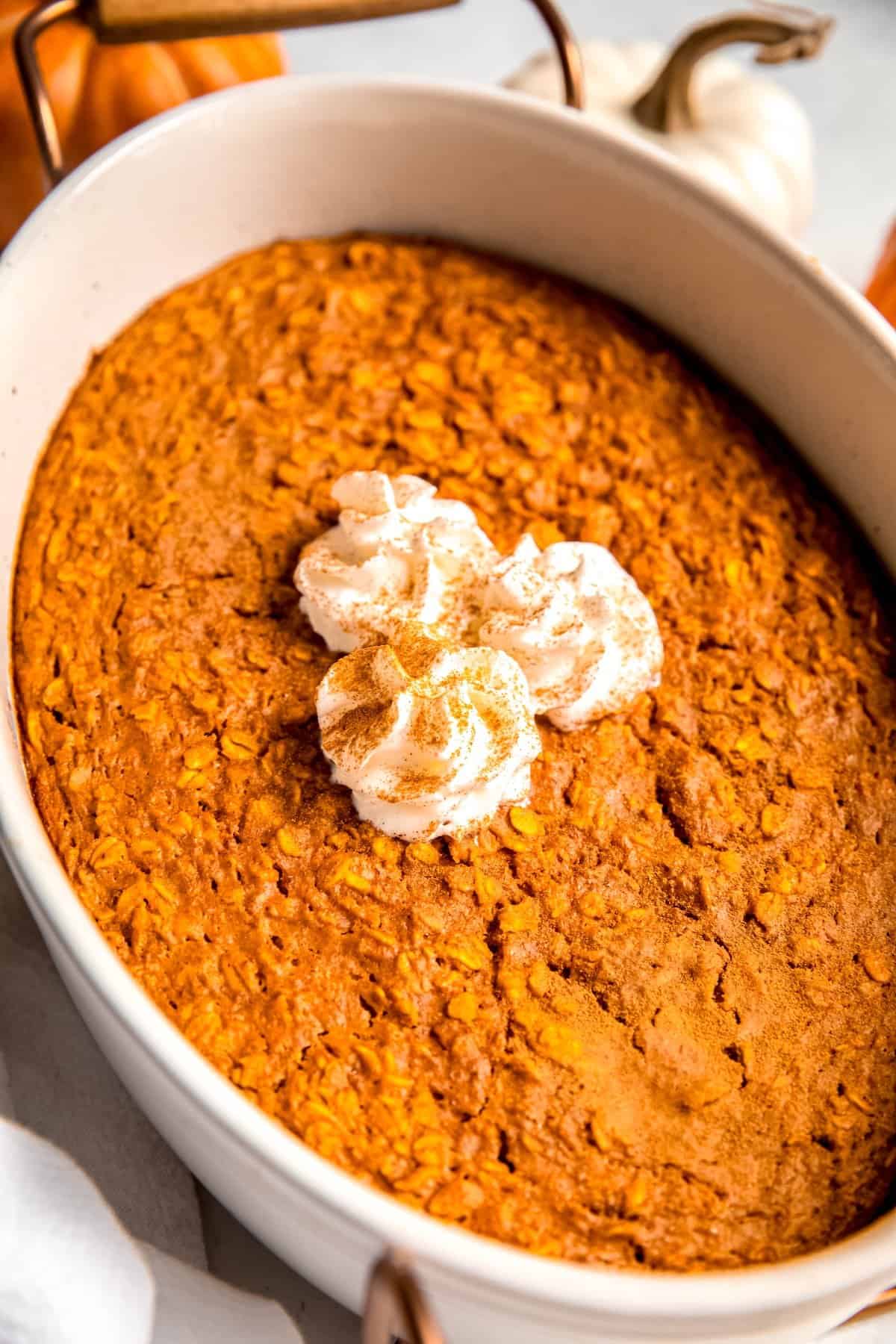 45 degree angle downward shot into a casserole dish filled with pumpkin pie baked oatmeal topped with 3 rosettes of whipped cream and a light sprinkling of whipped cream.