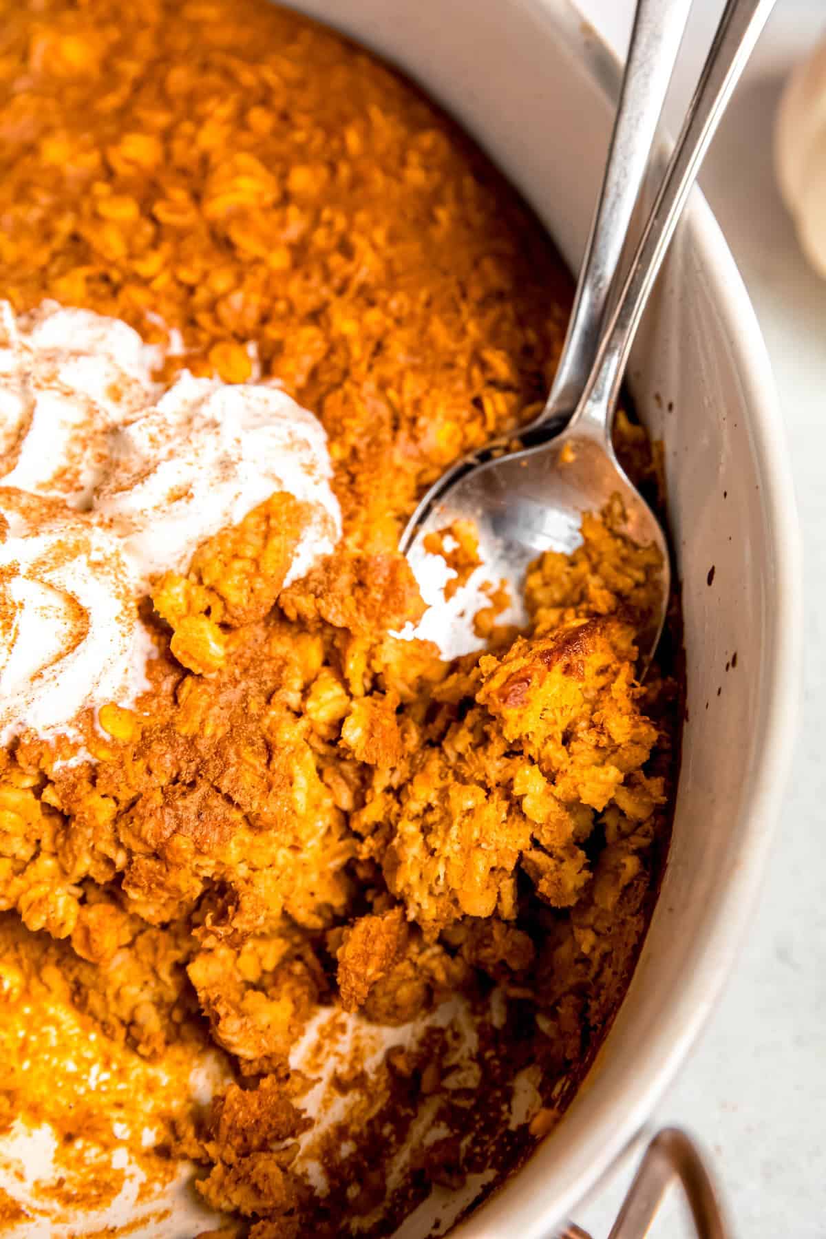 closeup shot of the casserole dish of pumpkin pie baked oats with two silver serving spoons that have been used to serve out several portions already.