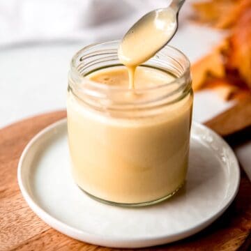 square hero image of creamy maple dijon salad dressing in a small glass jar on a white plate on a wooden cutting board with a silver spoon drizzling some into the jar to show the viscous consistency.