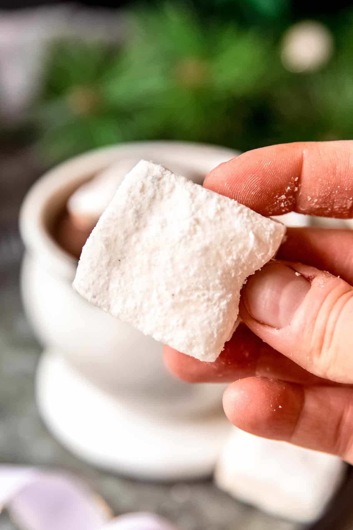 hand holding a dairy free marshmallow with the thumb pressing in a side to show the springy consistency; there's a mug of hot chocolate blurred out in the background.