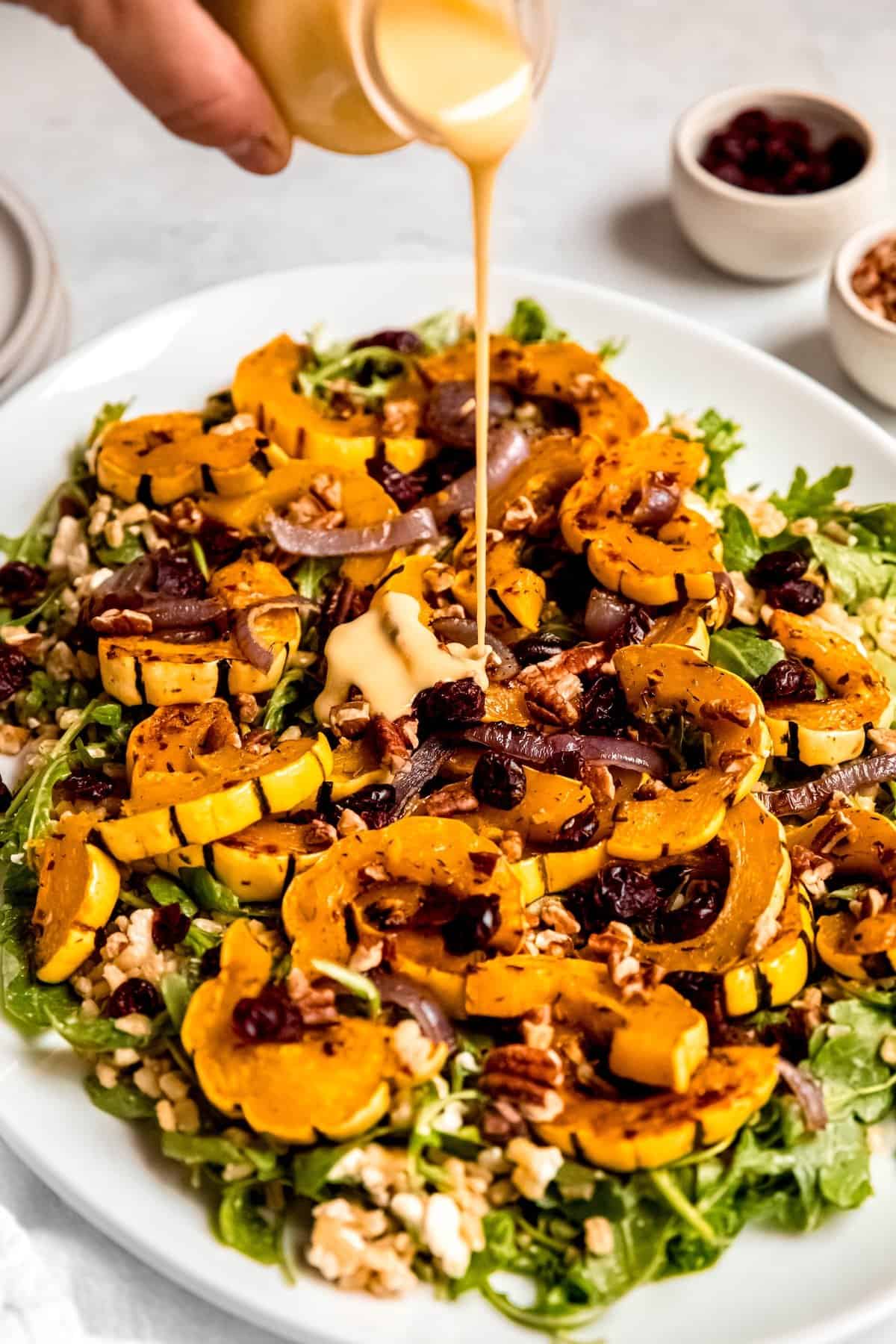 action shot of a hand pouring creamy maple dijon dressing onto a serving platter with roasted delicata squash and arugula salad.