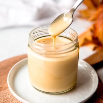 action shot of a hand holding a silver spoon drizzling some of the dijon maple dressing into a small glass jar showing how thick and creamy (but still pourable) it is.