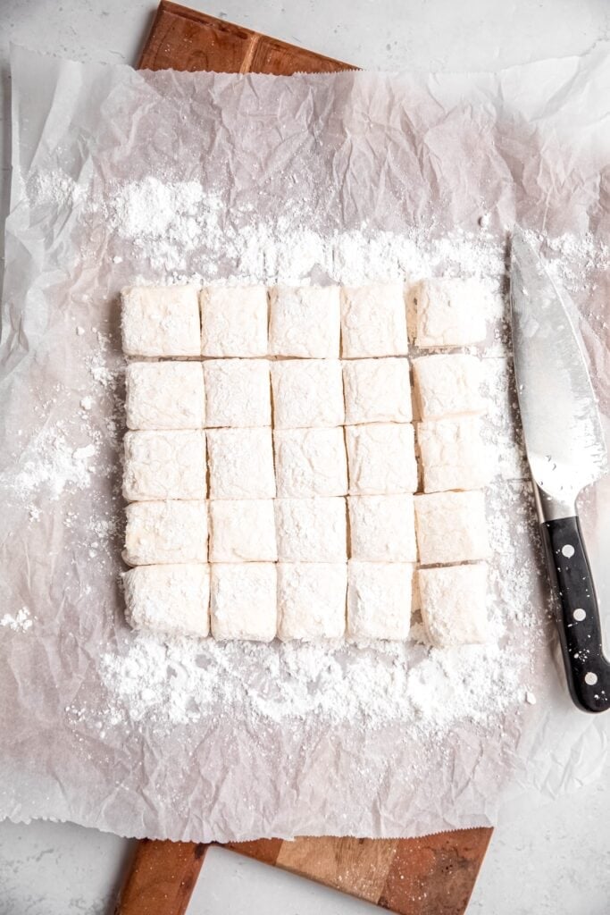 homemade egg white marshmallows on a piece of parchment on a wooden cutting board after cutting into 25 pieces.
