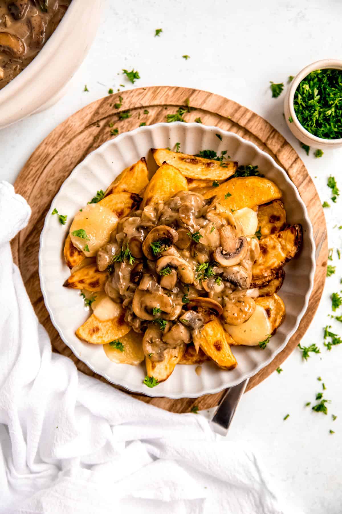 flat lay shot of a white scalloped bowl filled with a serving of poutine for breakfast on a white table with parsley scattered around.
