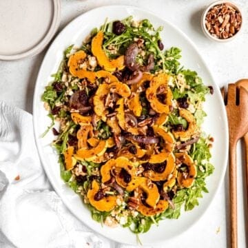square hero shot of a white oval serving platter with roasted delicata squash salad with dried cranberries, red onions, arugula, barley, pecans, and feta on a white table next to wooden serving spoons and a pinch bowl of pecans.