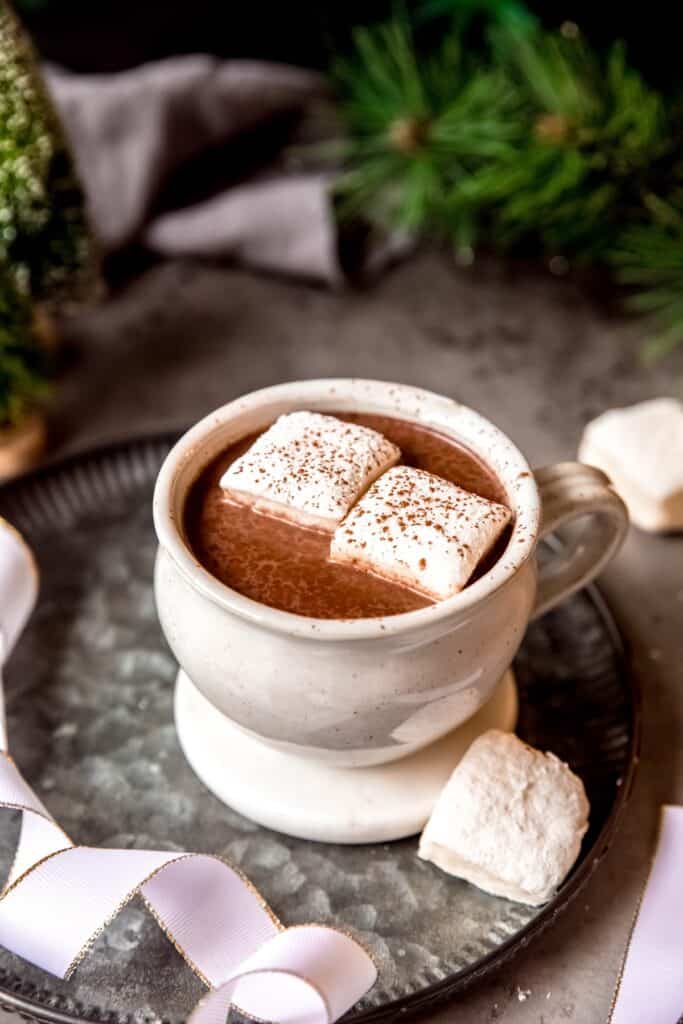 dark and moody wintry table with a white mug of dark aztec hot chocolate topped with white marshmallows and dark red chili powder with a gold-edged white ribbon and two marshmallow squares framing the mug with the dark pine and grey linen in the background.