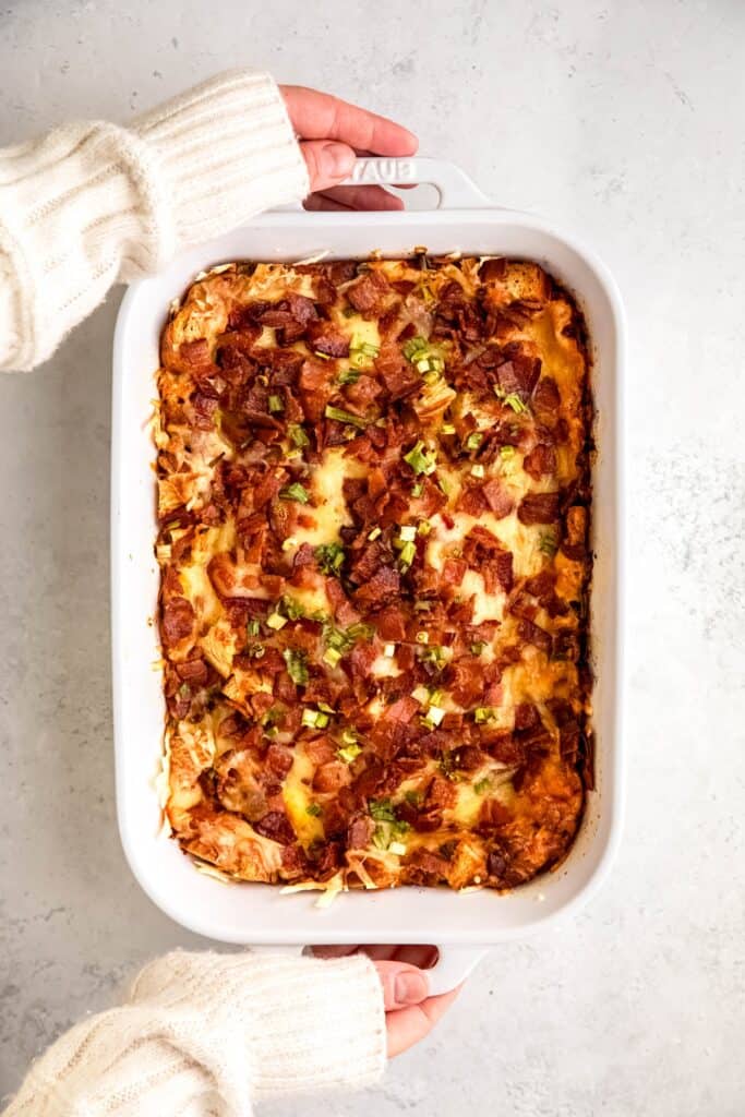 two hands in a white sweater up to the knuckles setting down a 9x13 of bacon swiss croissant breakfast casserole down on the table to cool.