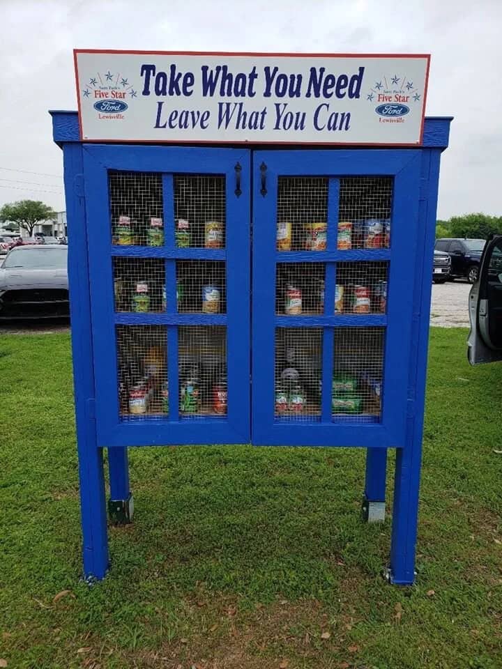 blue wooden hutch with casters turned into a community pantry with a big sign on top that says "take what you need leave what you can."