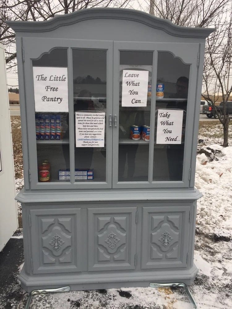 old armoire with glass front panels turned into a free pantry with signs taped on the inside saying "the little free pantry," "leave what you can," take what you need," and smaller instructions that aren't legible without squinting.