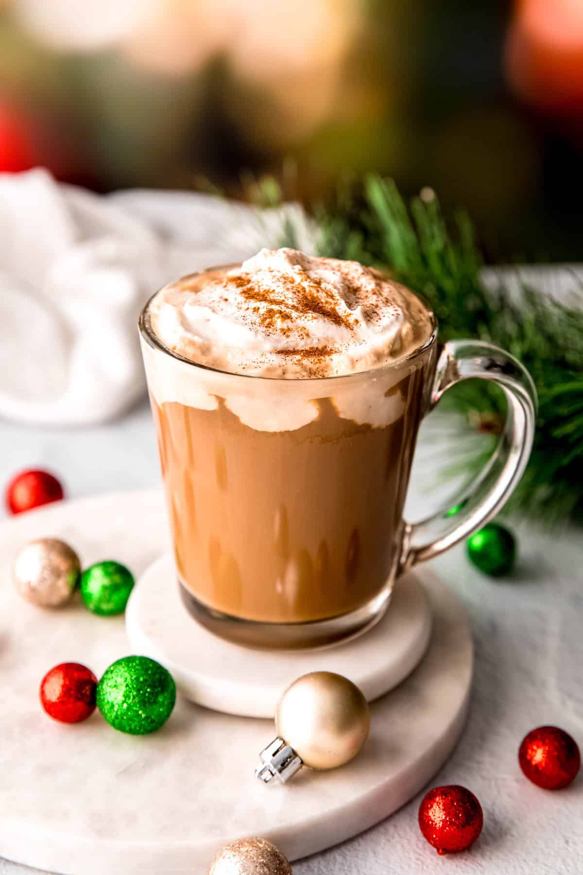 boozy christmas coffee in a clear glass mug topped with cinnamon on a white table; the mug is surrounded by varying sizes of small red, white and green holiday ornaments, a sprig of pine, and a blurred out lit christmas tree in the back.