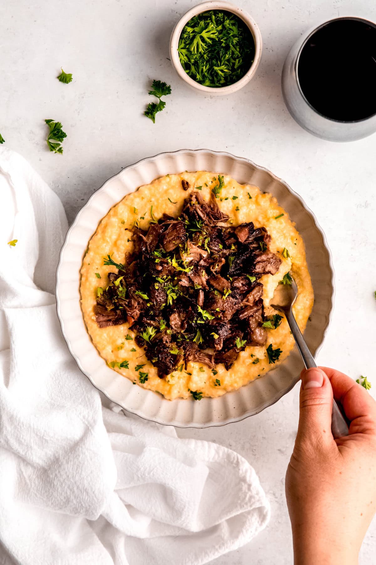 hand taking a forkful of wine braised beef short ribs and cheesy polenta from a bowl with a glass of red wine to the top right of the frame.