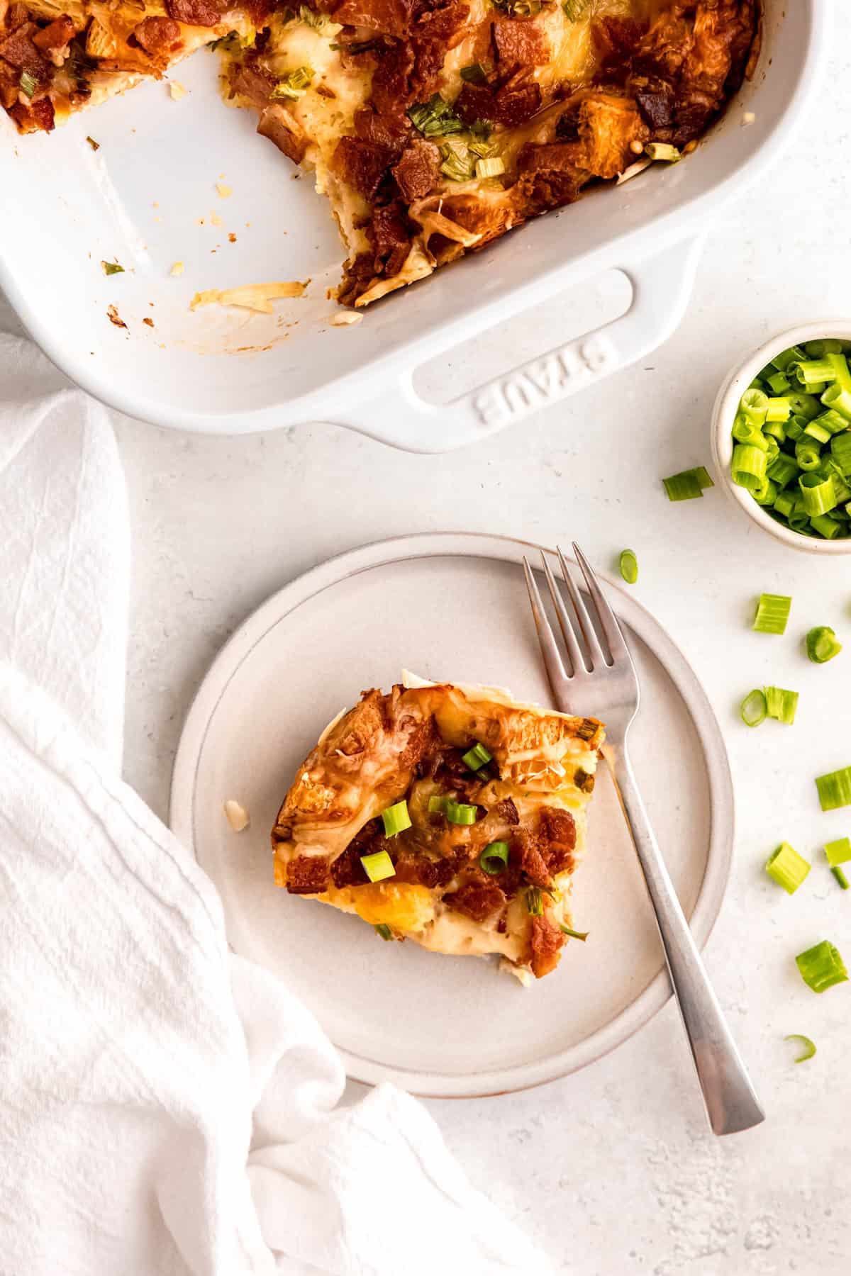 bacon and swiss croissant breakfast casserole in a 9x13 casserole dish with one slice removed and plated on a round white plate with extra scallions on top.