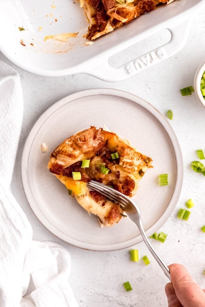 hand using a silver fork to cut the side of a slice of croissant bacon swiss strata with the rest of the breakfast casserole in the baking dish at the top of the frame.