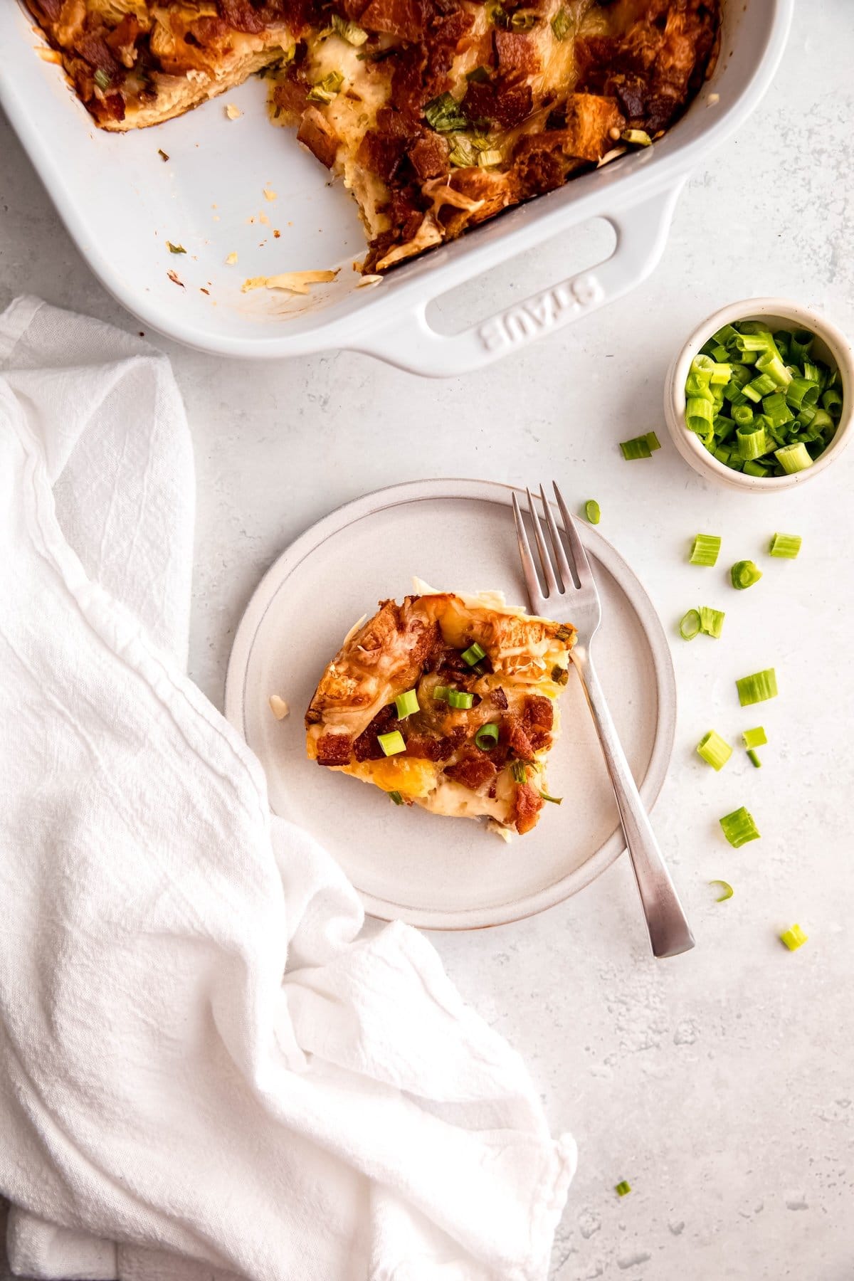 flat lay shot of a white table with a round white plate with a corner piece of bacon swiss croissant breakfast casserole with a silver fork, a pinch bowl of sliced scallions with some of the greens scattered around, and the rest of the casserole dish in the top of the frame.