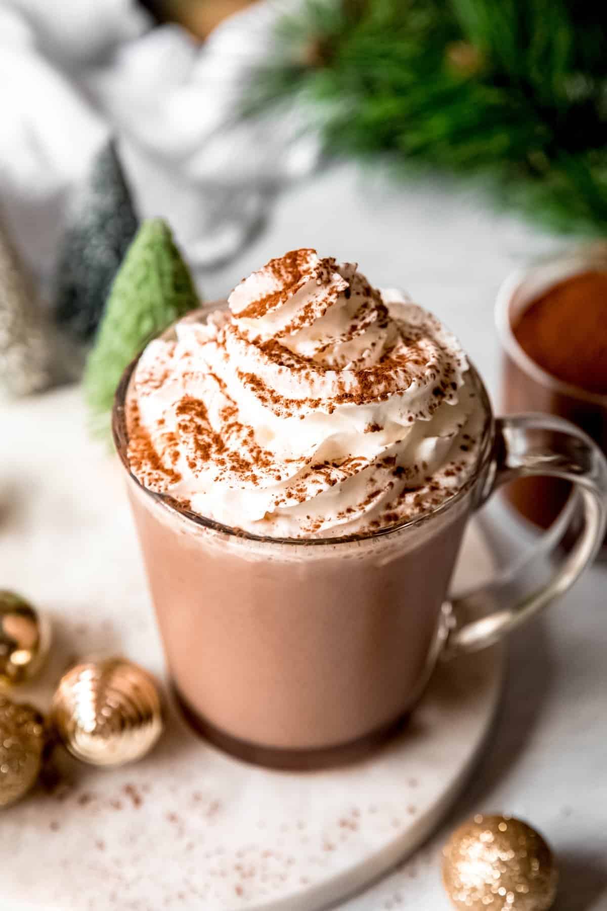 mug of whipped cream topped dark hot chocolate made from dark chocolate hot cocoa mix on a table with a sprig of pine, bottle brush trees, golden ornaments, and a glass with the rest of the homemade cocoa mix.