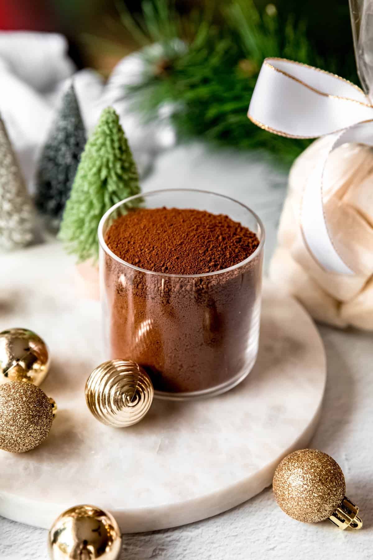 jar of dark hot chocolate mix on a table with christmas ornaments, bottle brush trees, and a cellophane bag of homemade marshmallows wrapped with a white and gold ribbon.