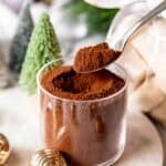 silver spoon taking a spoonful of dark hot chocolate mix from a clear glass jar.