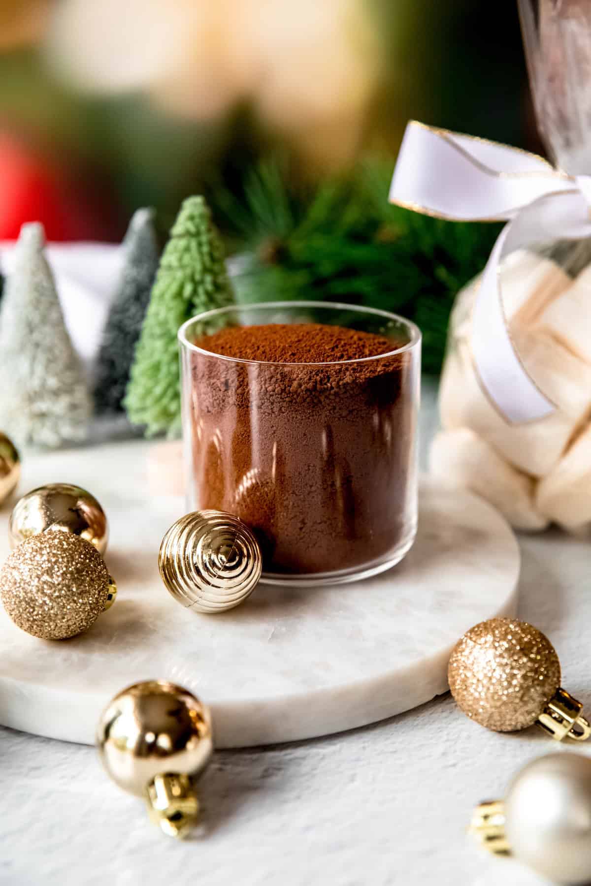 jar of dark hot cocoa mix with a bag of homemade marshmallows as a homemade christmas gift on a table with christmas lights, bottle brush trees, and small gold ornaments.