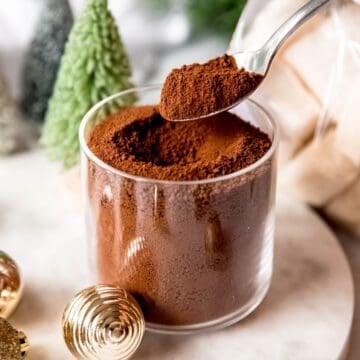 square hero image of a silver spoon dipping into a clear glass of dark hot cocoa mix to show the fine consistency of the homemade powder.