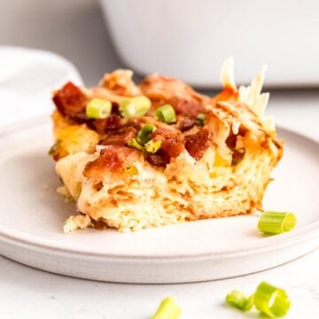slice of bacon swiss breakfast strata showing the fluffy croissant layer topped with cheese, meat, and scallions with the rest of the casserole dish blurred out behind.