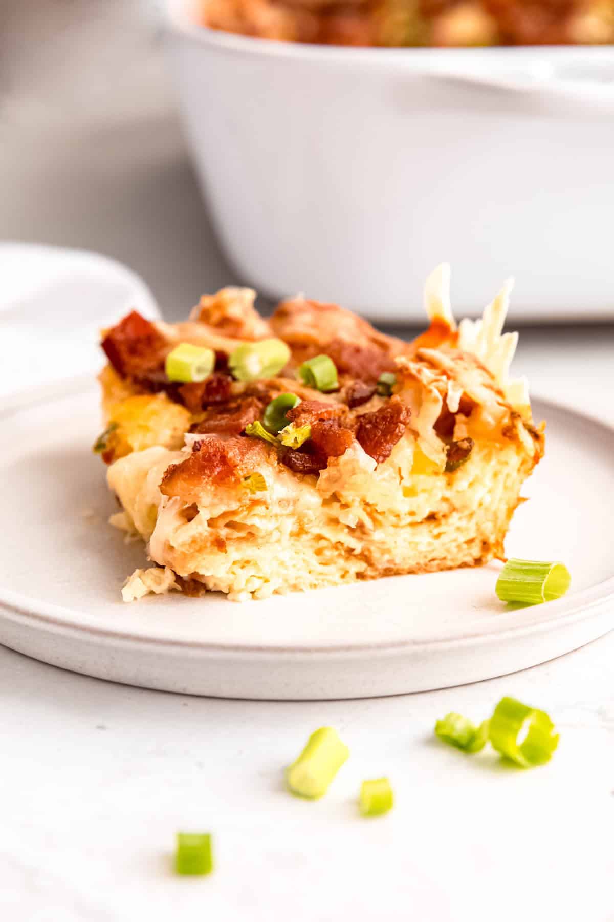 slice of bacon swiss breakfast strata showing the fluffy croissant layer topped with cheese, meat, and scallions with the rest of the casserole dish blurred out behind.