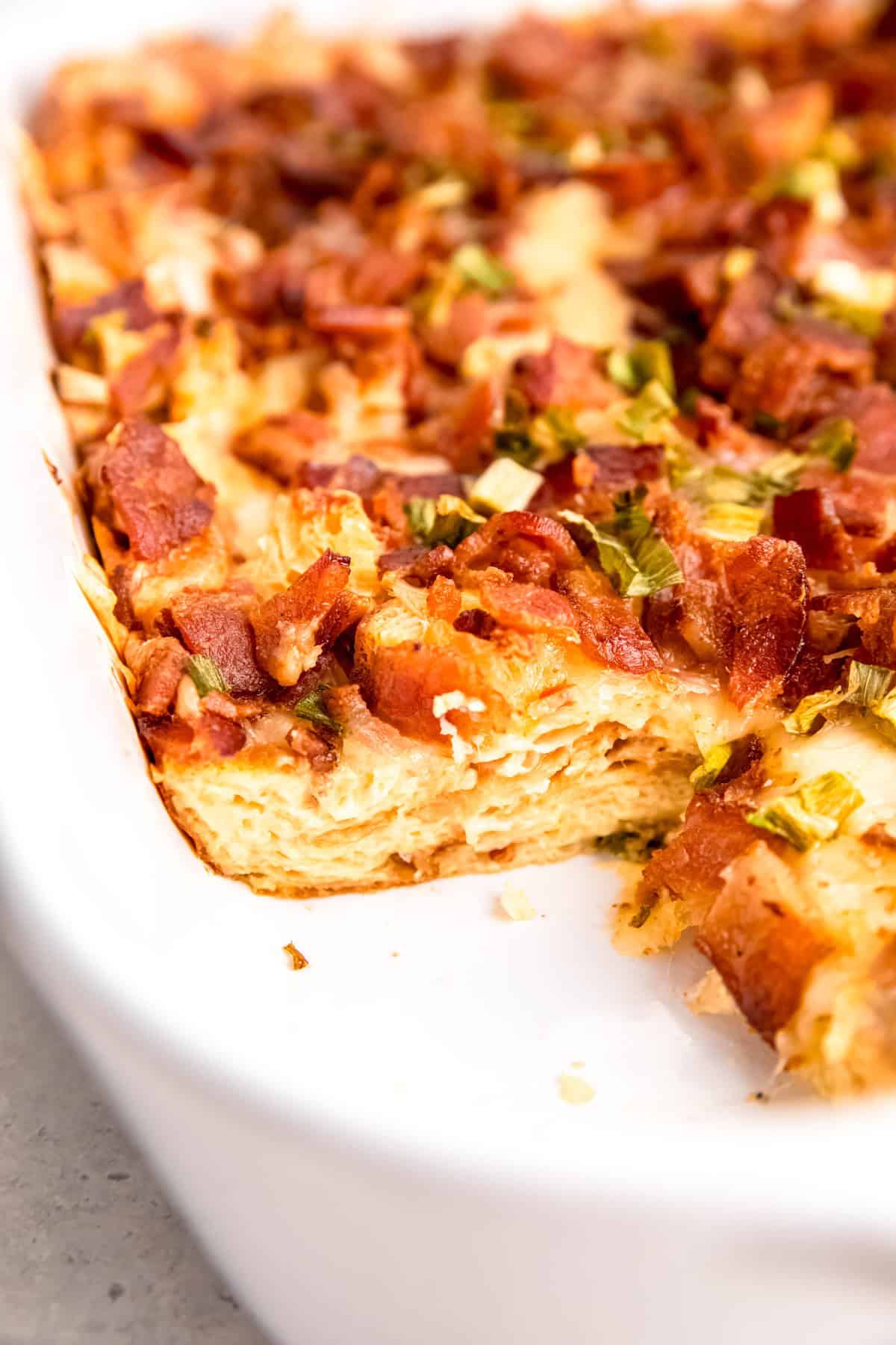closeup on the inside corner of a casserole dish with a fluffy make ahead croissant strata with the corner piece taken out; you can see the layers of croissant soaked in custard on the bottom, melty cheese, crispy bacon, and green onions on top.