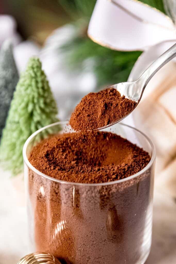 action shot of homemade dark hot cocoa mix being sprinkled into a clear glass jar from a silver spoon; you can see the fine grit of the mixture as it falls.