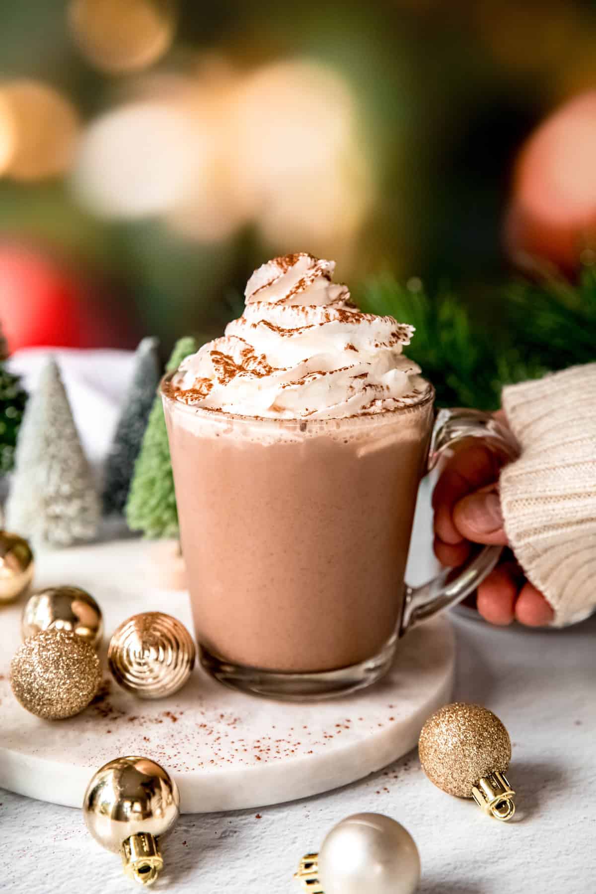 hand grabbing a clear mug of homemade dark hot cocoa topped with whipped cream and chocolate shavings from a white table with bottle brush trees, gold ornaments, and a pine sprig in front of a blurred out wall of christmas lights.