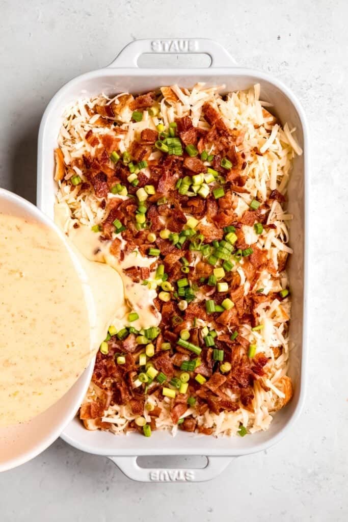 action shot of the egg custard mixture being poured over the assembled bacon swiss strata.