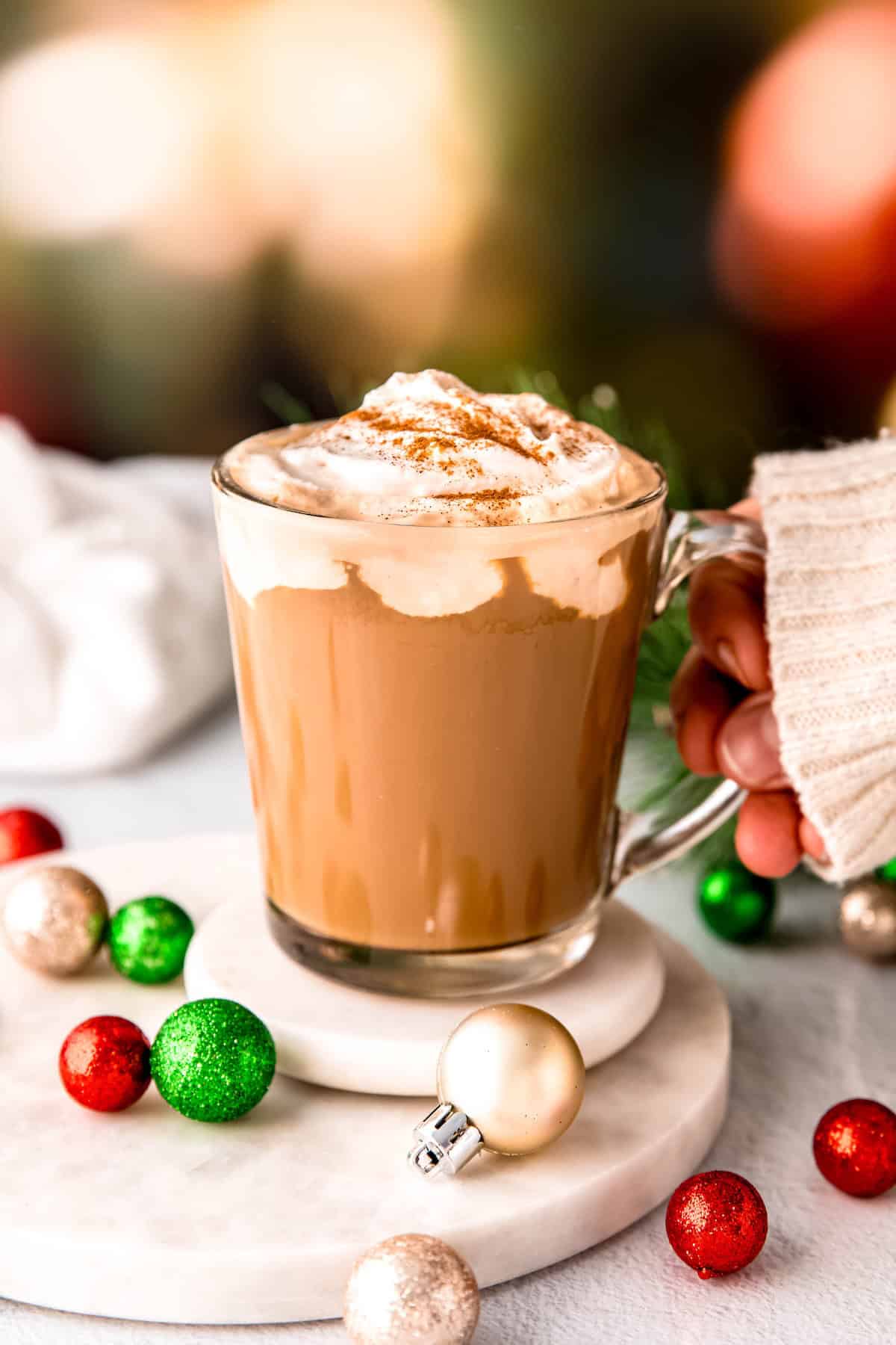 hand in a white sweater up to the knuckles grabbing the handle of a clear mug filled with boozy christmas coffee from a white table with holiday ornaments.