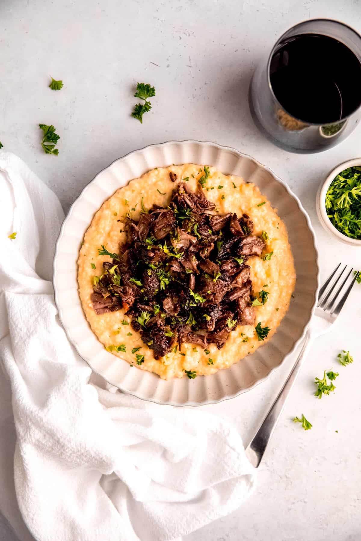 overhead shot of a round white pasta bowl filled with a serving of red wine braised short ribs on a bed of creamy parmesan polenta on a white table with a silver fork, a glass of red wine, a pinch bowl of fresh parsley, and a white linen napkin.