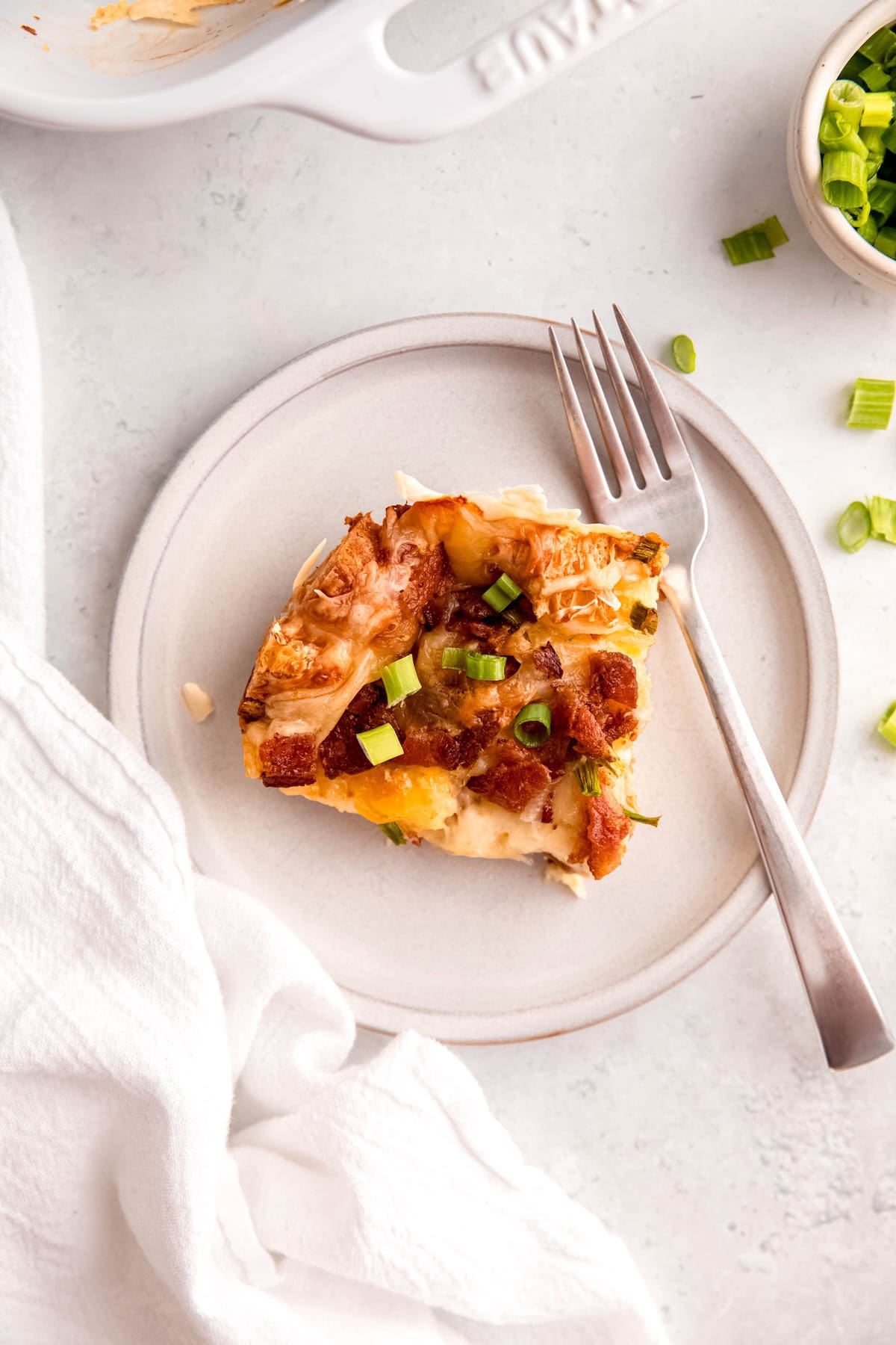overhead shot of a slice of make ahead croissant breakfast casserole with bacon, swiss and green onions on a round white plate with a silver fork.
